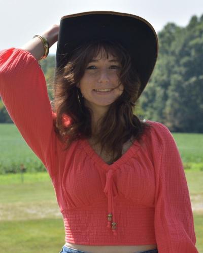 A woman in a red top and black hat smiles at the camera, with her hand on her hat.