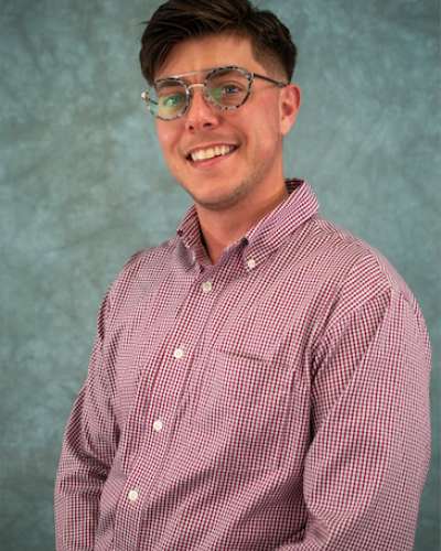 A man in a red and white checkered shirt and glasses smiles while looking at the camera.