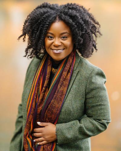 A woman with curly hair, wearing a green blazer and colorful scarf, smiles while standing in front of a blurred background.