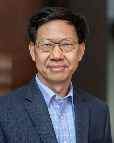 A man wearing a blue suit jacket and glasses smiles while looking at the camera.