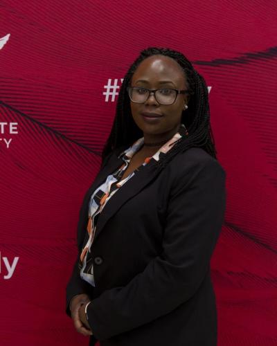 A woman stands in front of a red backdrop, wearing a black blazer and a patterned blouse.