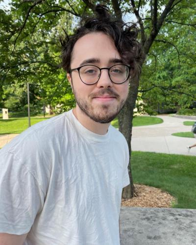 A man wearing a white t-shirt and black-framed glasses smiles at the camera in front of a tree and landscaped area.
