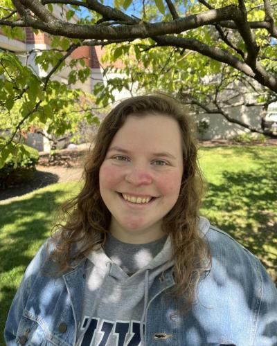 A young woman with long brown hair smiles while wearing a denim jacket and gray hoodie outside under a tree.